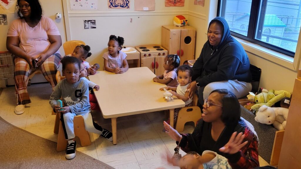 A group of parenting adults and children sit in an early childhood classroom during a Play with Purpose playgroup session. Two adults are singing to the group.