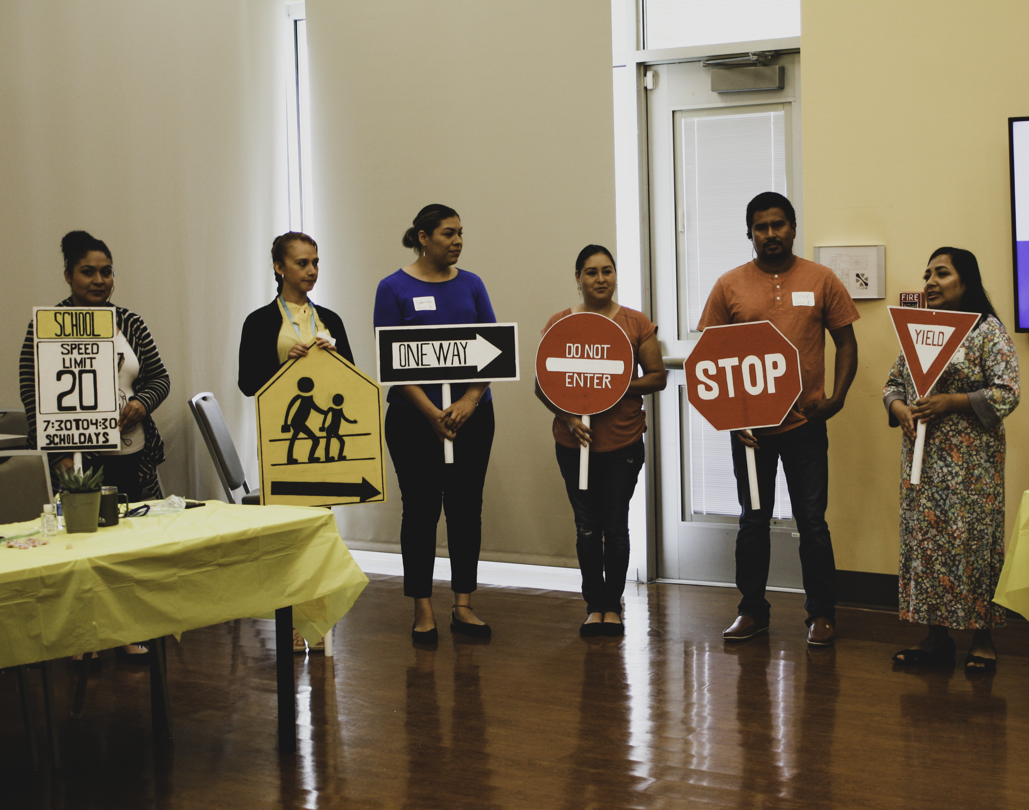 A group of participants of Activate! Local are standing in a line at the front of a meeting room. Each holds a traffic sign and one person is speaking.